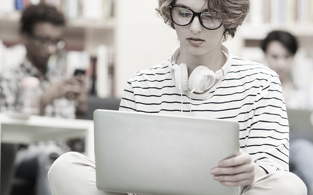 A young person looking at a computer, a young man behind them with a phone and another with a computer.