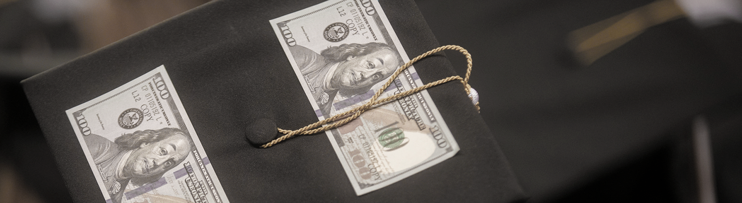 Part of a graduation cap with two 100 dollar bills pinned to it.