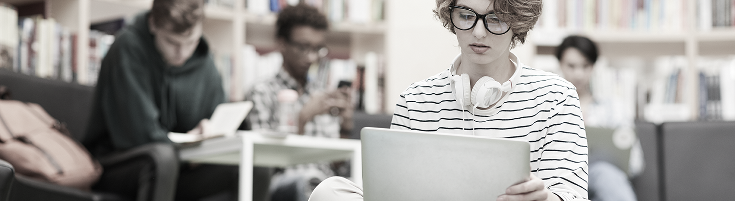 Students in a library, person with curly hair, wearing headphones around neck and looking at a computer. 